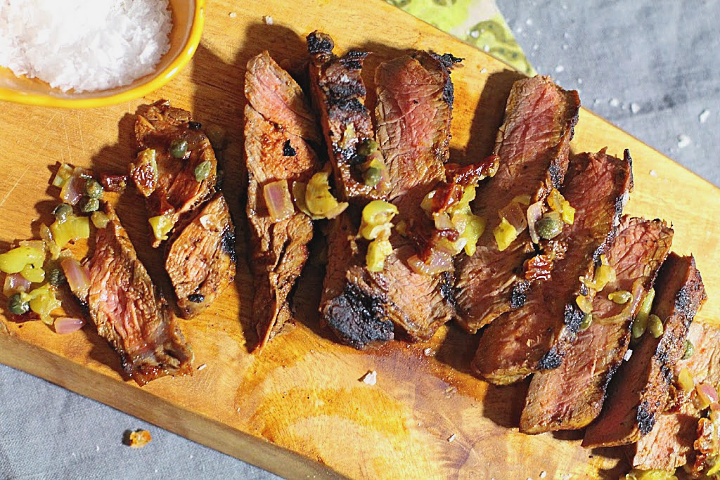 Sirloin steak pan grilled and sliced on a cutting board. Topped with pepperoncini, caper, dijon, sun-dried tomato relish