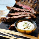 Marinated flank steak on a black wood cutting board with a bowl of horseradish cream sauce