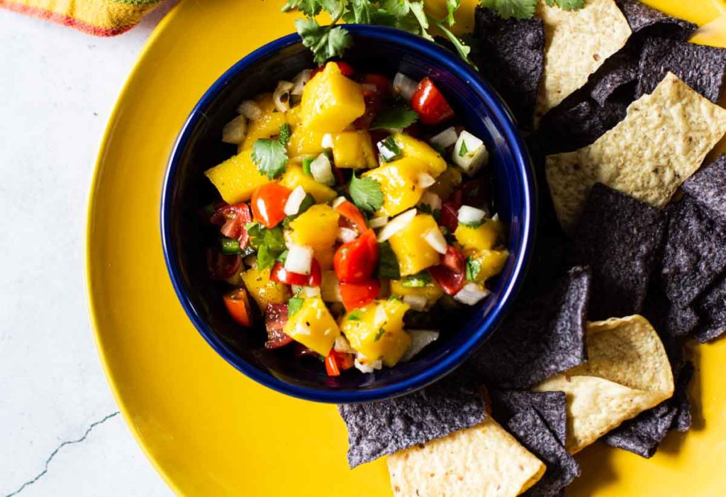 Mango salsa in a blue Fiesta Bowl on a yellow plate with tortilla chips.