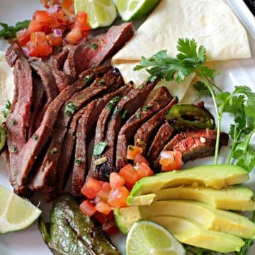 A platter of carne asada with avocado, lime, tomatoes and a flour tortilla