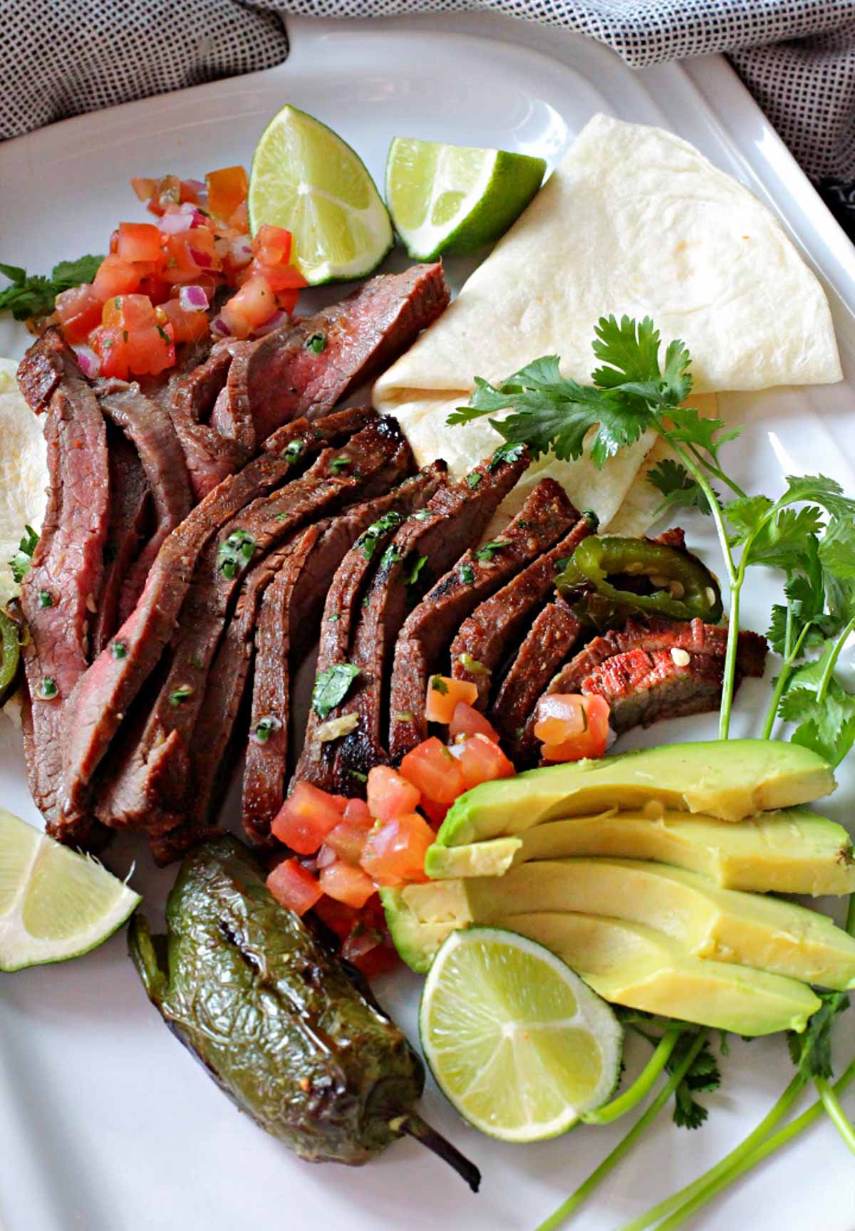 A platter of carne asada with avocado, lime, tomatoes and a flour tortilla