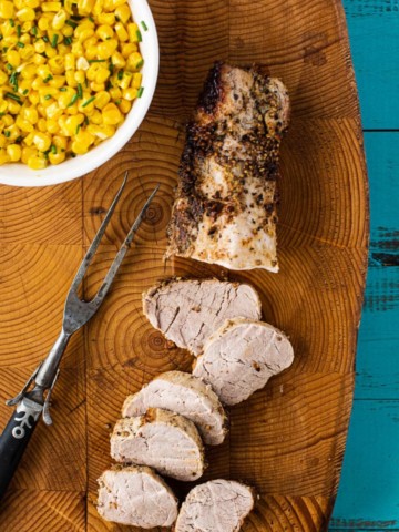Baked pork tenderloin coated with dijon mustard and coriander and peppercorns. Sliced on a cutting board served with fresh corn.