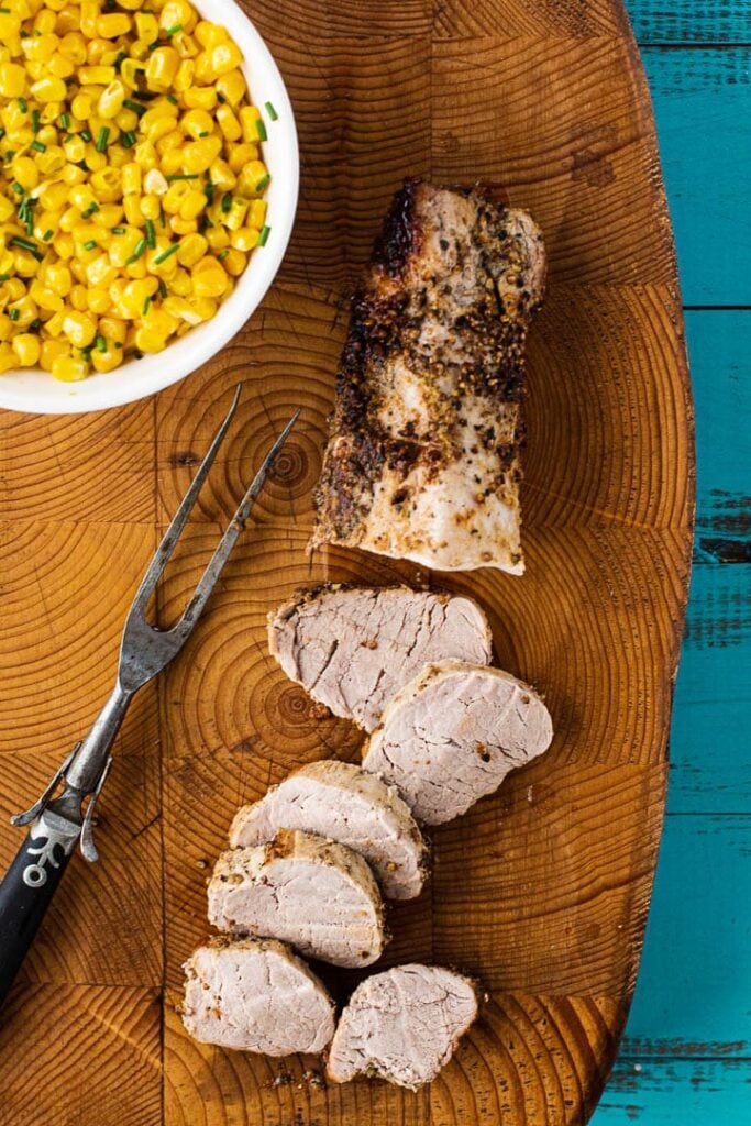 Baked pork tenderloin coated with dijon mustard and coriander and peppercorns. Sliced on a cutting board served with fresh corn.