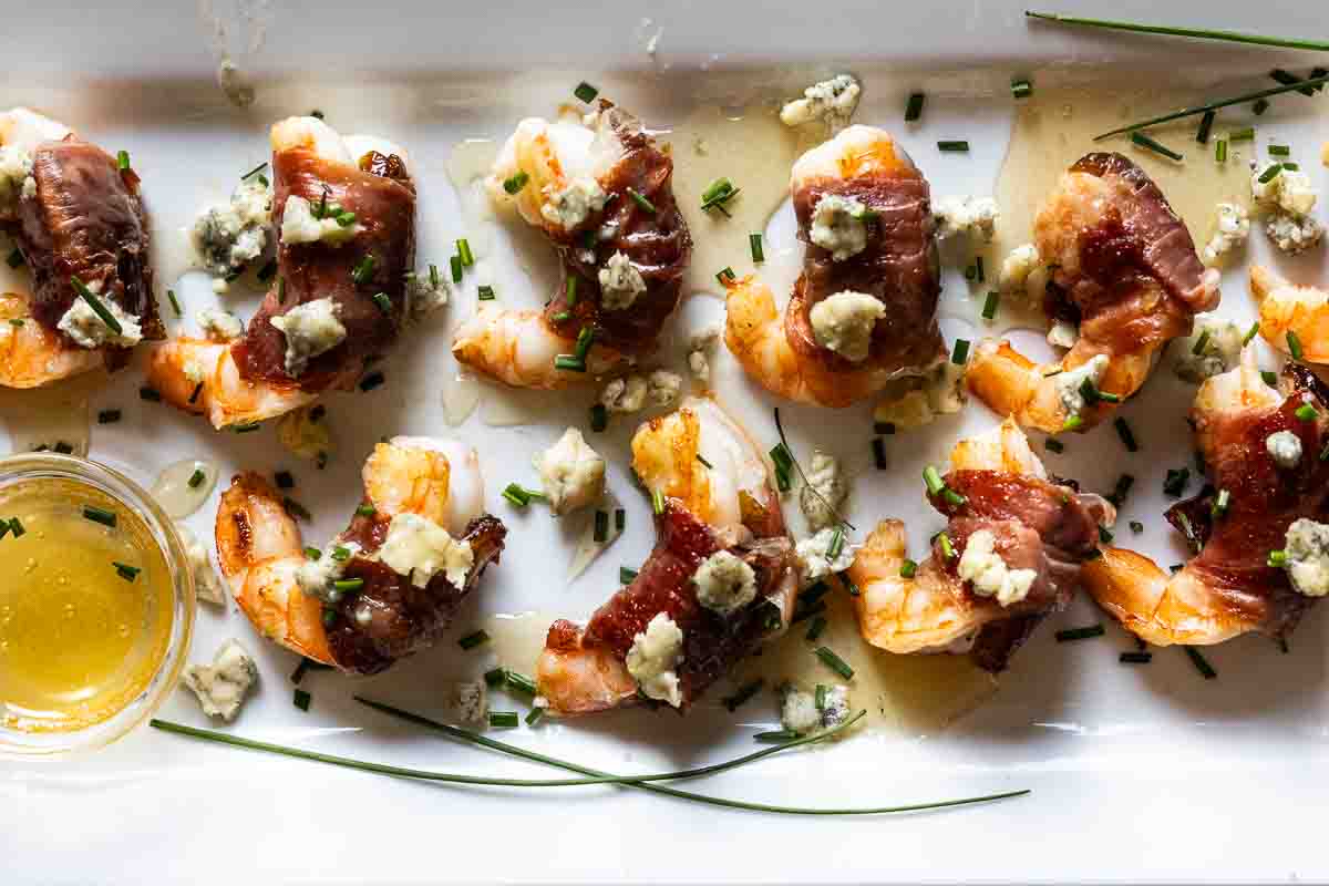 Prosciutto wrapped shrimp on a white platter drizzled with honey and garnished with chopped chives.