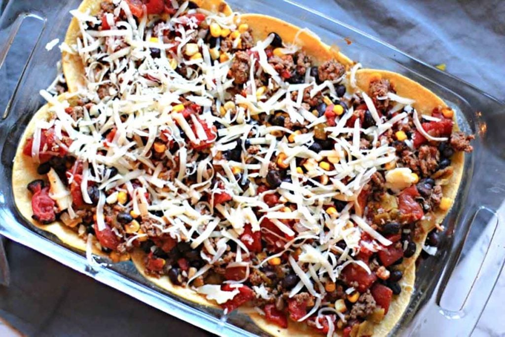 Mexican casserole layered with ground beef and corn tortillas topped with cheese, ready for oven