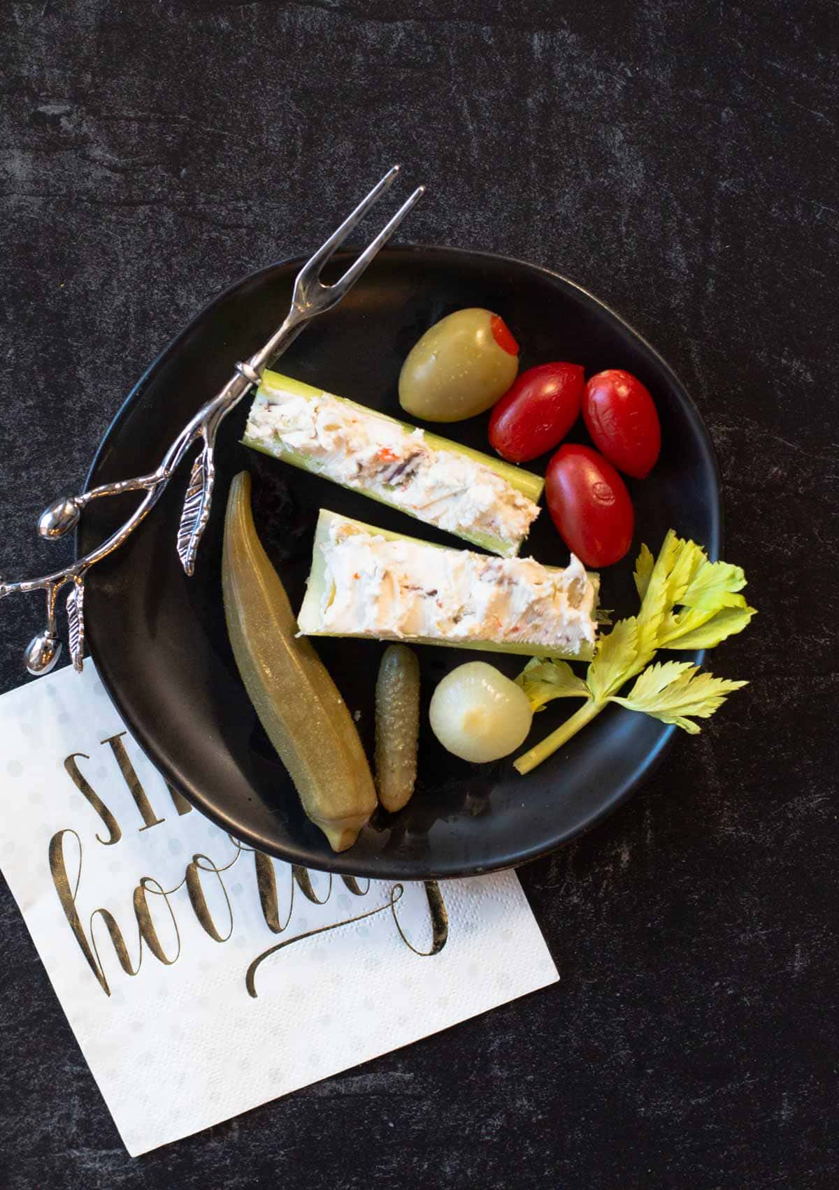 A black appetizer plate with cream cheese stuffed celery sticks, cherry tomatoes, and pickled okra