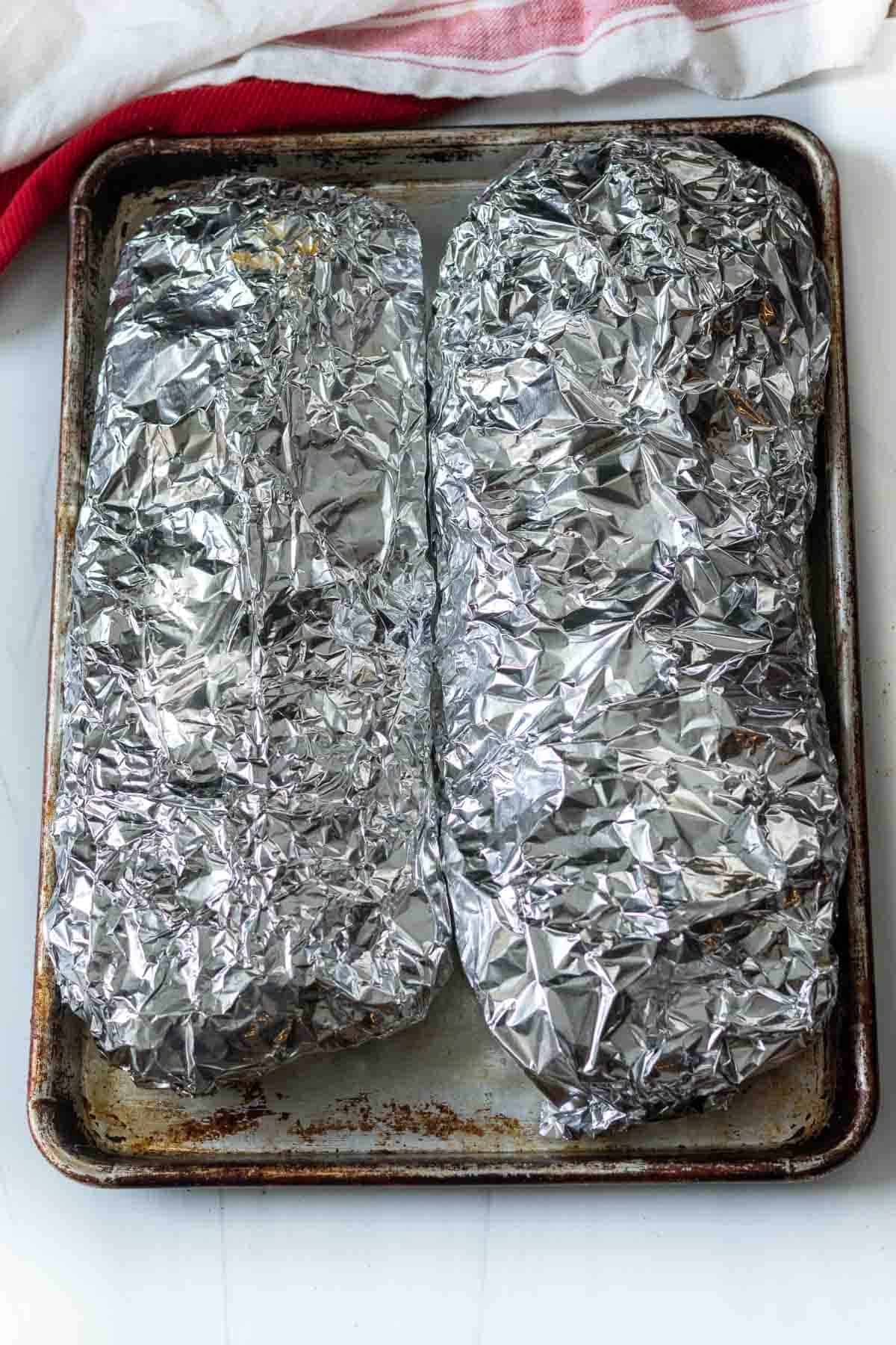 Two racks of ribs wrapped in foil and on a sheet pan ready to bake.