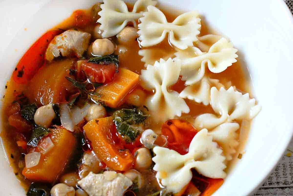 Butternut squash soup with chicken, chickpeas and bowtie pasta in a white bowl.