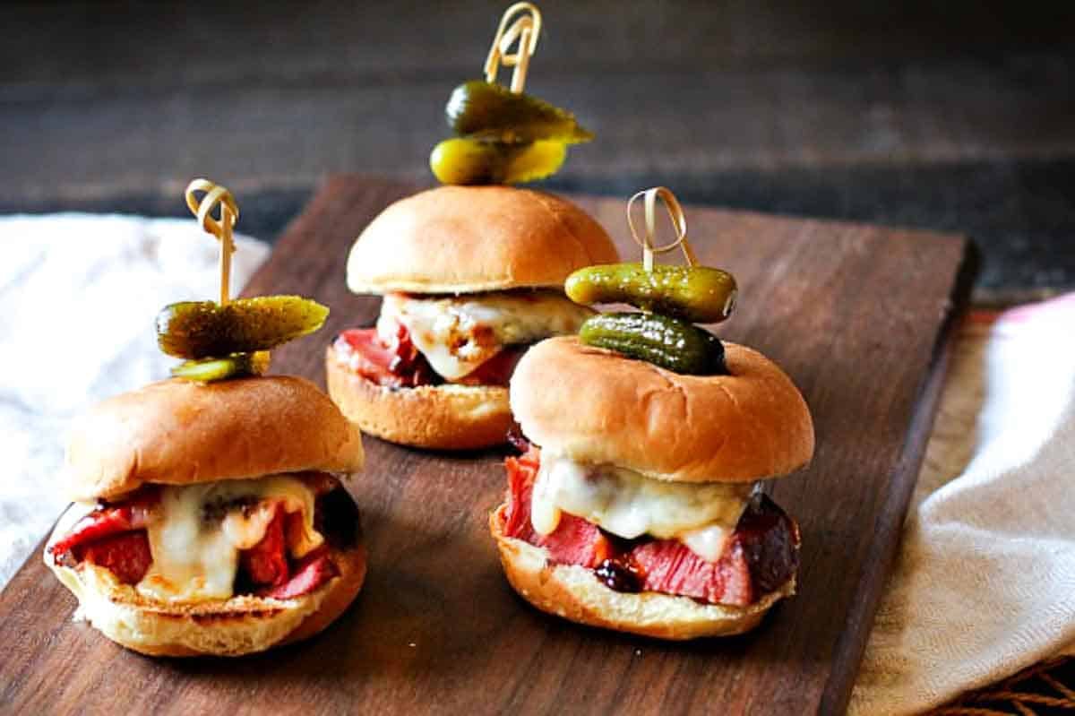 Glazed corned been sliders on a wood cutting board garnished with pickles.