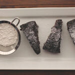 Chocolate banana bread recipe cut in wedges, sprinkled with powdered sugar on a rectangular white serving platter