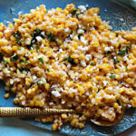 Mexican corn salad sprinkled with Mexican Crumbling Cheese and cilantro. Served on a blue hand made ceramic platter with a gold spoon.