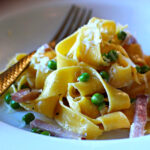 Pappardelle pasta with peas and ham and lemon cream sauce served in a white pasta bowl with a gold fork.