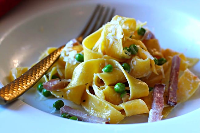 Pappardelle pasta with peas and ham and lemon cream sauce served in a white pasta bowl with a gold fork.