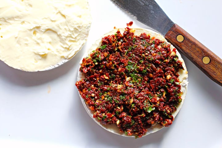 A mixture of sun dried tomatoes and basil spread on half of a wheel of brie cheese.