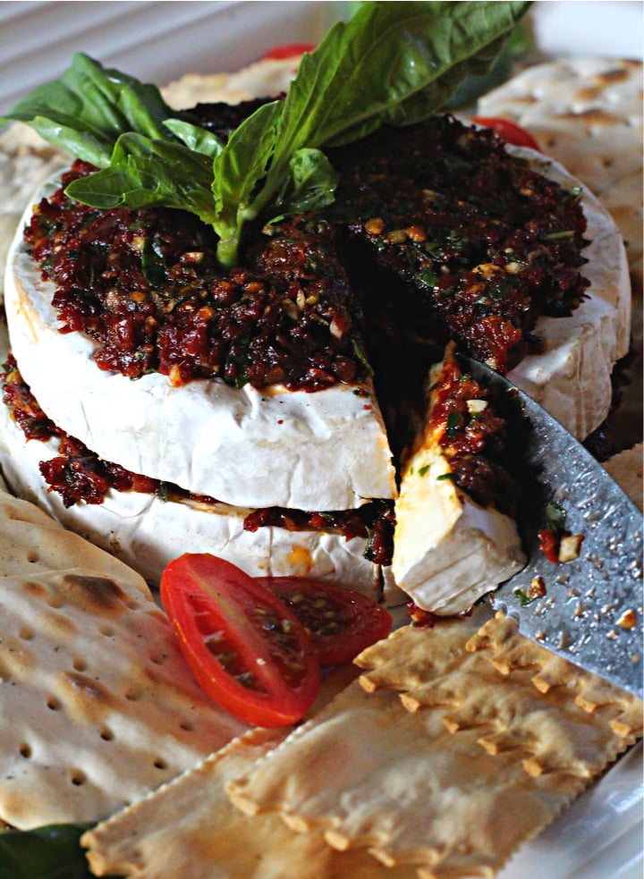 Two halves of a wheel of brie cheese topped with sun-dried tomatoes in oil and served with water crackers and garnished with fresh basil leaves