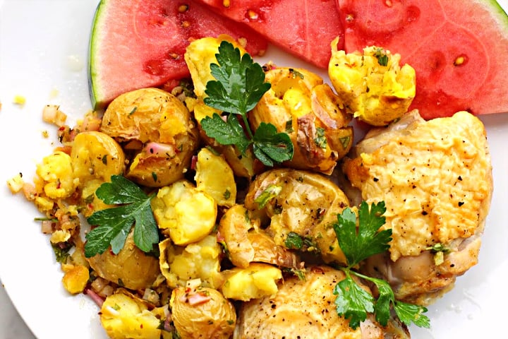 Smashed potatoes topped with chimichurri sauce next to baked chicken thighs and slices of watermelon