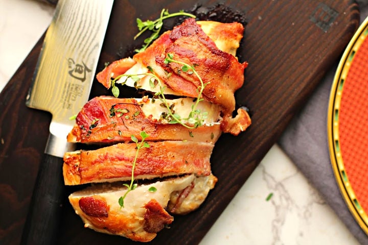 bacon wrapped chicken breast with springs of thyme on a wood cutting board sliced with a Japanese knife.
