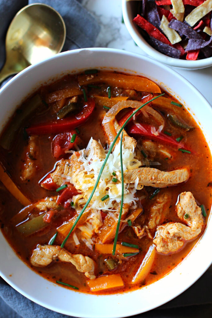 Chicken fajita soup topped with grated Mexican cheese, served in a white soup bowl and a gold soup with. with a bowl full of tortilla strips for garnish.Topped with fresh chives.