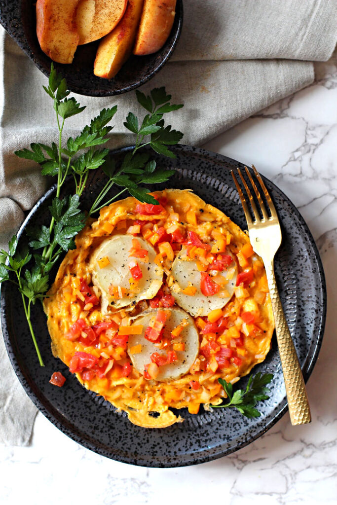 Espagnole omelette served on a black plate, sprinkled with tomatoes, onion and orange bell peppers and three slices of potato.