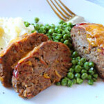 Individual spicy meatloaf on a plate with green peas and mashed potatoes.