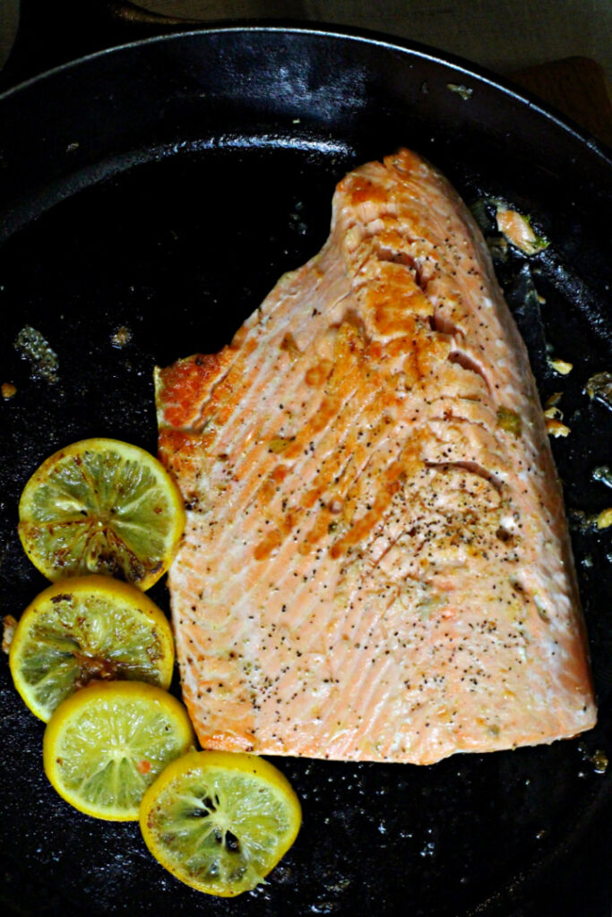 A salmon fillet cooking in a cast iron skillet surrounded by lemon slices.