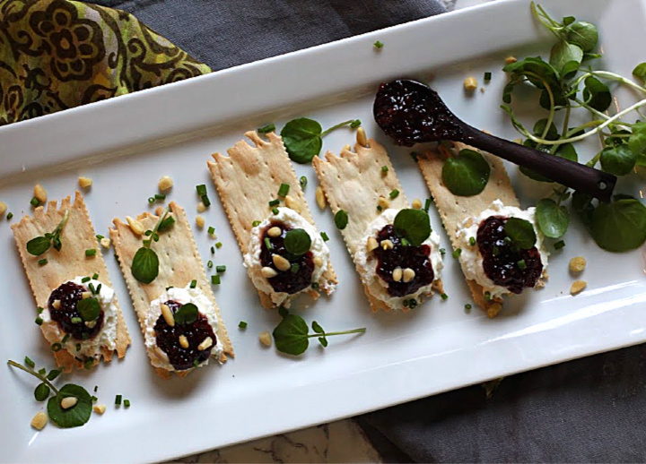 Wafer crackers topped with whipped goat cheese and topped with Heidi's Red Chile Raspberry Jam. Garnished with pinon nuts, chives and watercress. On a white serving platter