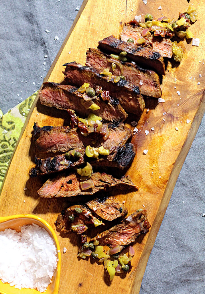 Sliced sirloin steak on a cutting board topped with pepperoncini and caper relish and sprinkled with Maldon sea salt.