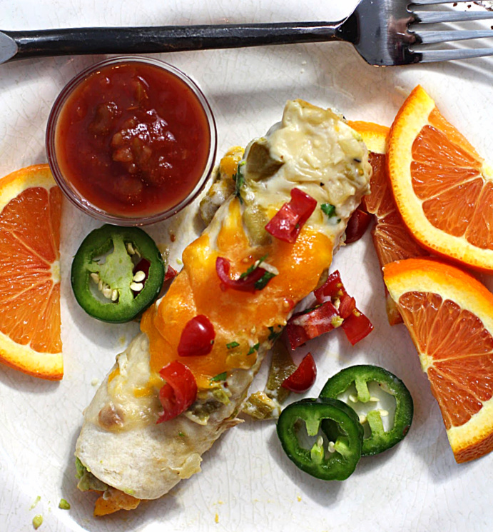 Smashed avocado breakfast burritos with scrambles eggs. Topped with melted cheddar cheese and garnished with slices of fresh jalapeno and chopped tomatoes. Served with salsa and sliced oranges.