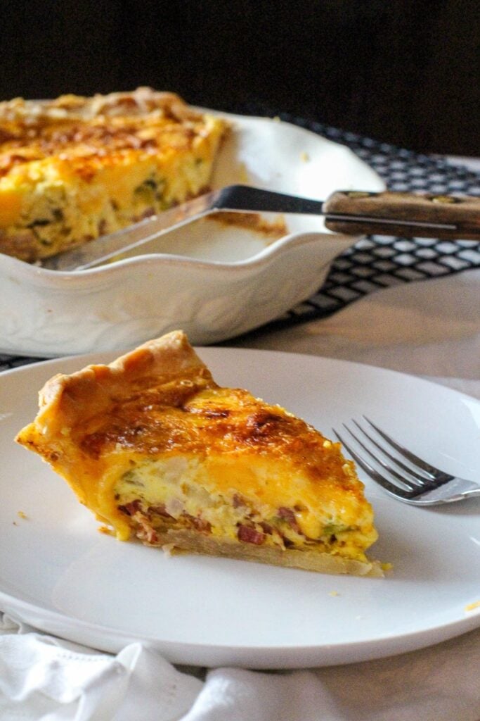 A slice of hatch green chile bacon quiche with a deep dish pie pan with quiche and spatula