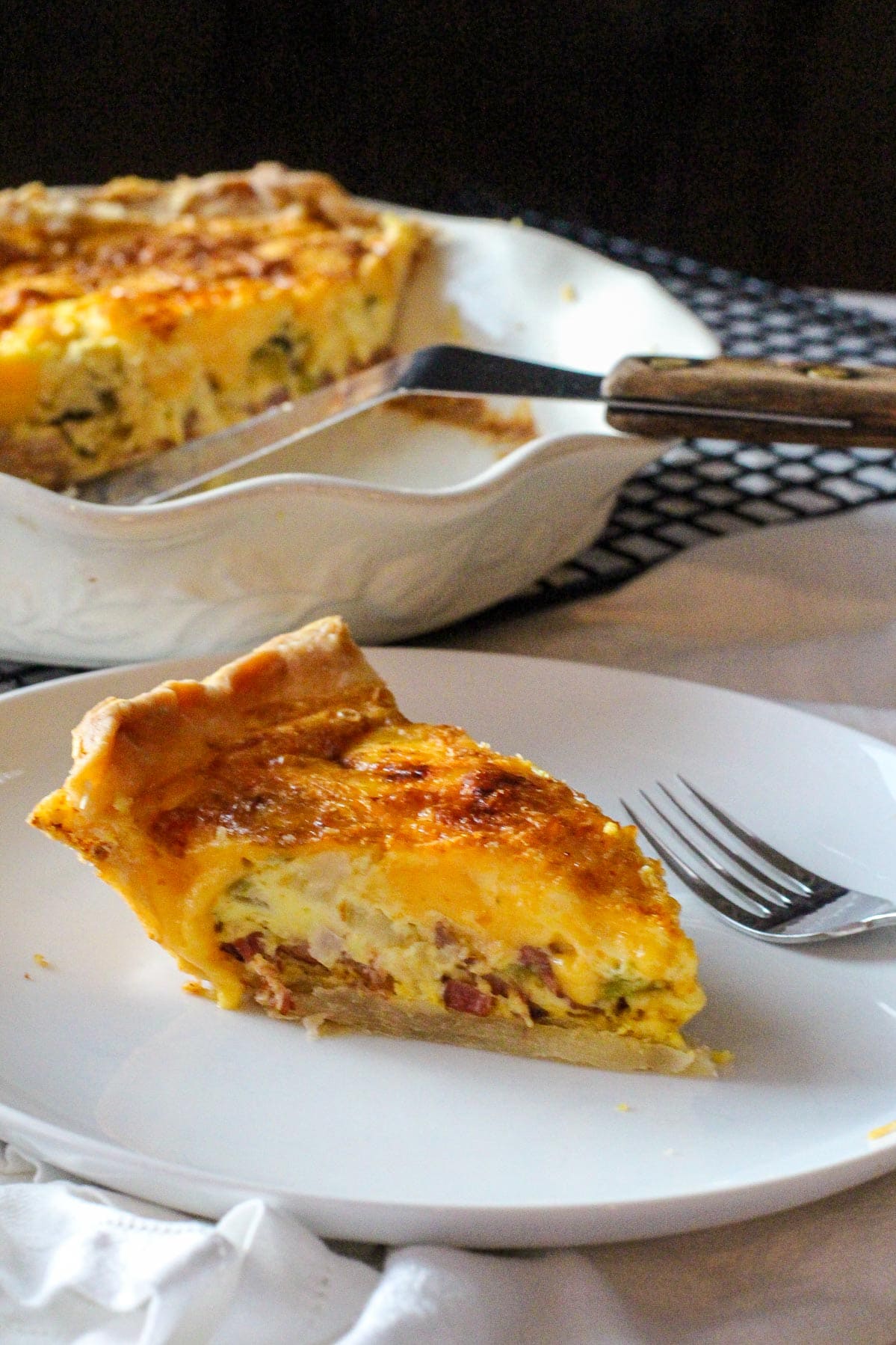 A slice of hatch green chile bacon quiche with a deep dish pie pan with quiche and spatula