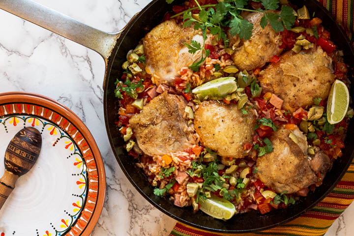 Arroz con Pollo, Mexican chicken and rice using chicken thighs cooked in a cast iron skillet garnished with lime wedges, cilantro and green olives.