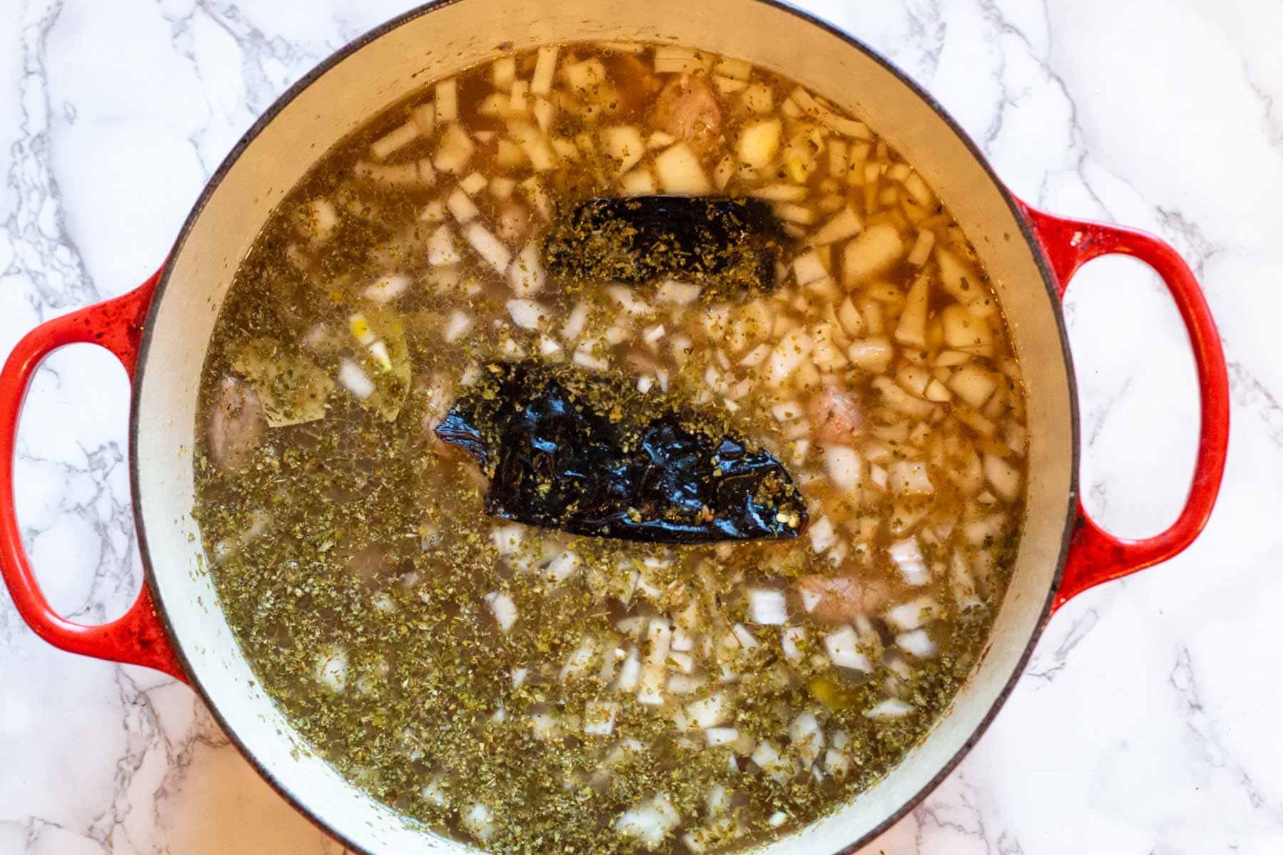 Adding spices and dried guijillo peppers to pork pozole ingredients in a dutch oven