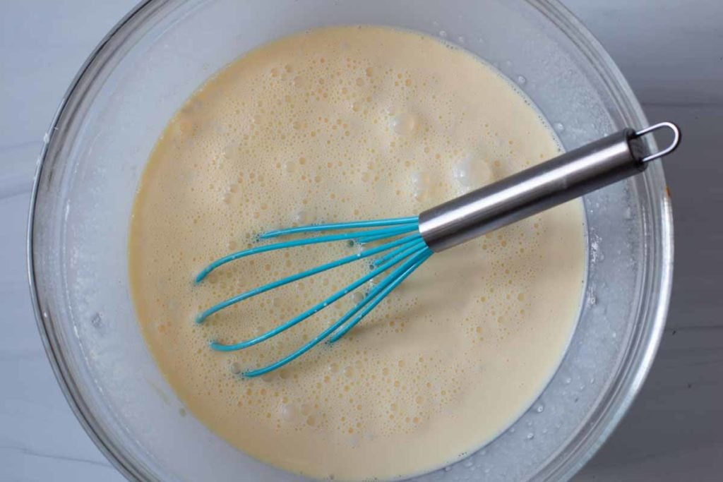 Egg, milk and flour mixture in a clear bowl with a blue whisk