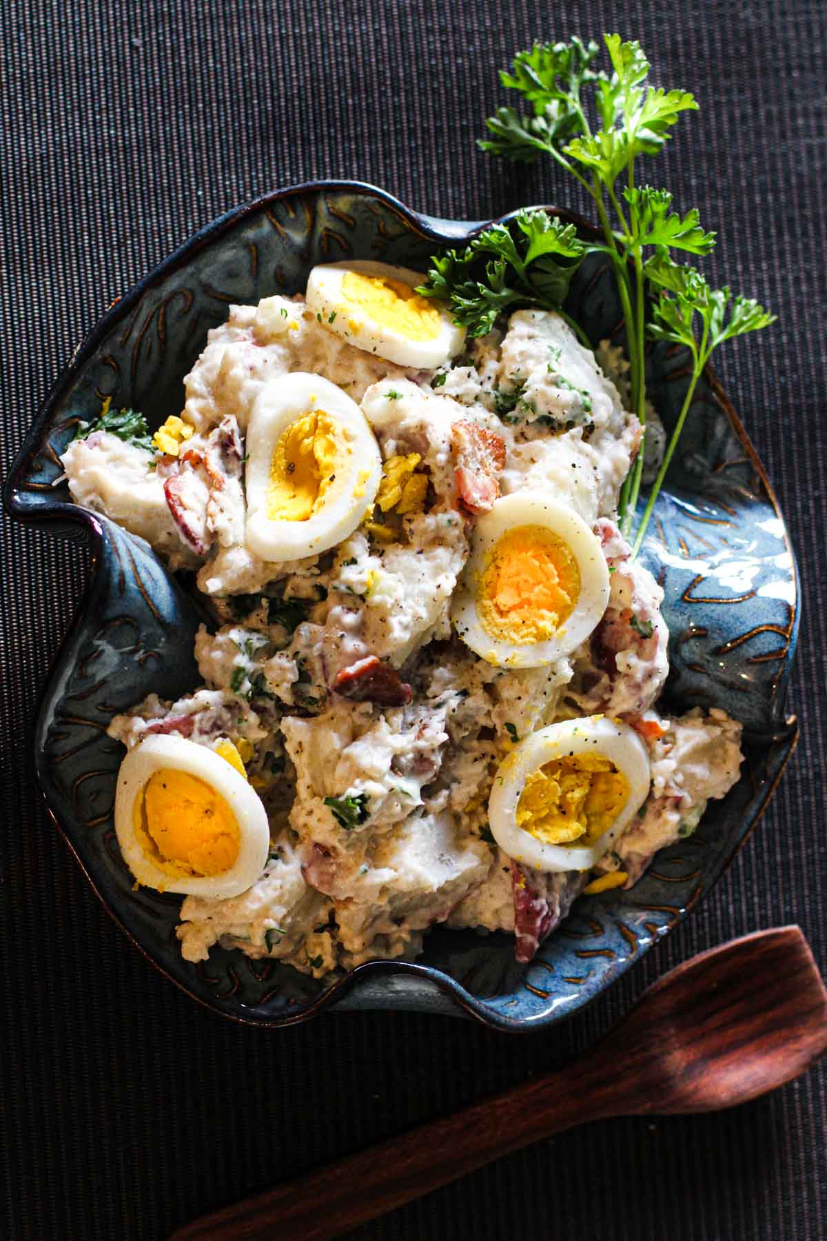 Blue cheese potato salad in a blue ceramic bowl topped with sliced hard boiled eggs and garnished with parsley