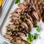 Sliced grilled Flank steak and chimichurri on a white platter.
