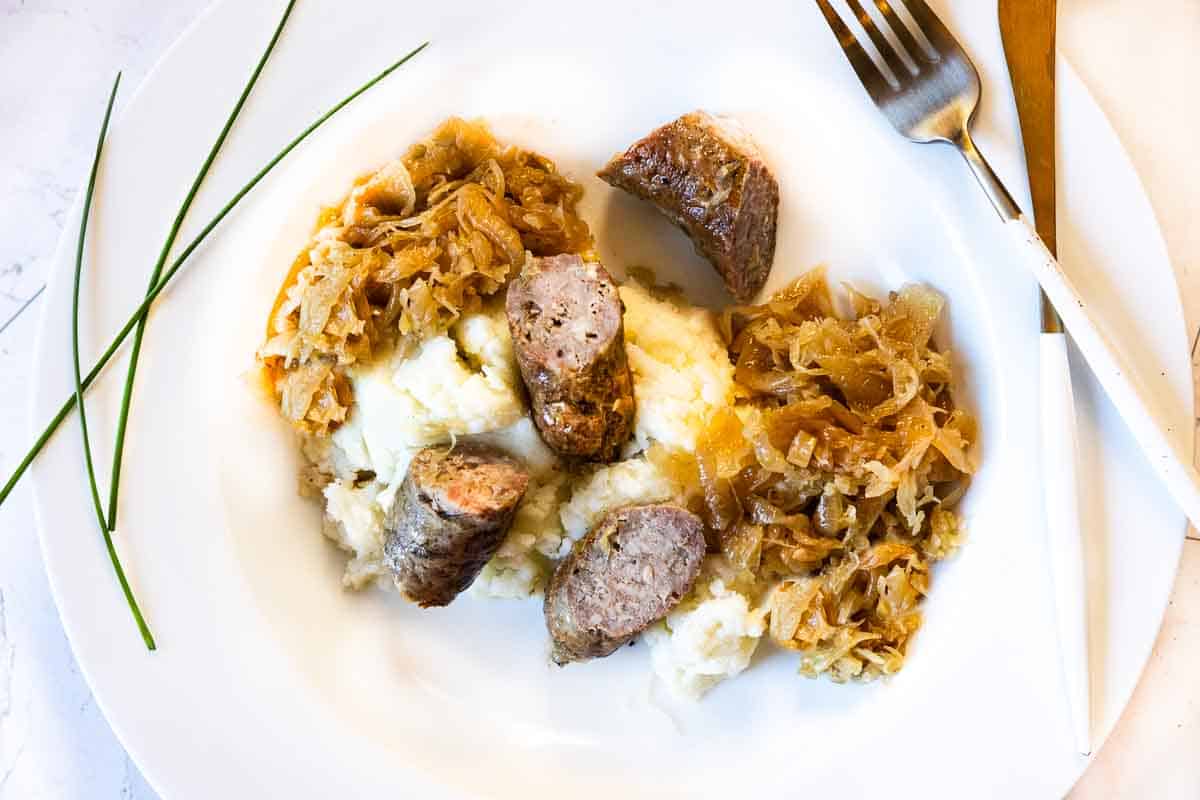 Brats and sauerkraut served over mashed potatoes on a white platter plate.