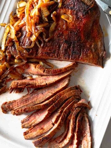 Sliced cast iron flank steak served on a white platter with charred onions.